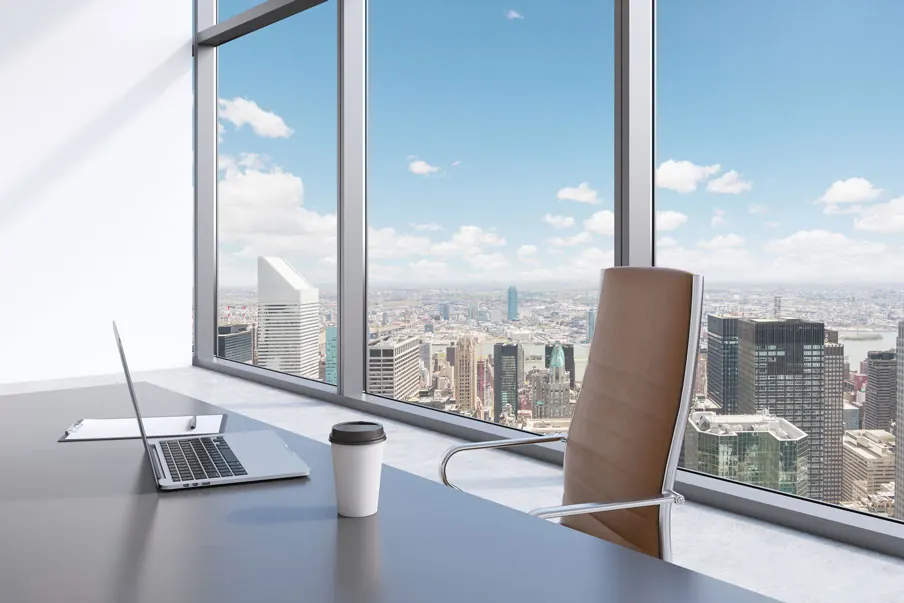 A quiet corner office: desk with a laptop and coffee, an empty leather chair, and the city beyond floor-to-ceiling windows.